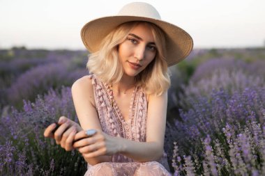Güzel bir kadın canlı bir lavanta tarlasında oturuyor, şık bir yaz elbisesi giymiş, şapkası var, dingin bir cazibe sergiliyor ve hafif açık hava aydınlatması altında mevsimlik moda sergiliyor..