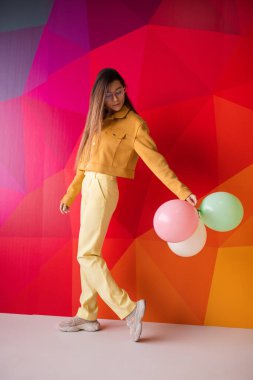 Sarı elbiseli şık bir genç kadın renkli bir geometrik arkaplan ve balonlarla ayakta duruyor. Titreşimli atmosfer neşe, tarz ve bireysellik yayar..