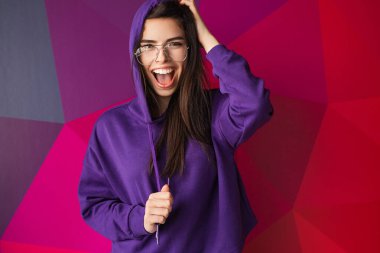 Mor kapüşonlu, neşeli genç bir kadın heyecanını dile getiriyor ve canlı, renkli bir geometrik arkaplana karşı duruyor. Pozitif enerjisi ve moda tarzı gençlik ve yaratıcılıkla yankılanıyor..