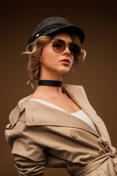 Элегантная женщина в бежевом пальто в солнцезащитных очках и черной шапке на нейтральном фоне, демонстрирующая современную моду с оттенком классического стиля.