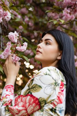 Pembe kiraz çiçeklerini, hayat dolu pınarları ve doğal güzelliği gözlemleyen bir kadının huzurlu görüntüsü. Yumuşak ışık ve çiçekli çevre sakin, taze ve düşünceli bir atmosfer yaratır..