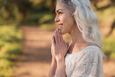 Gözleri kapalı ve elleri sımsıkı kapalı sakin bir kadın dışarıda duruyor, doğa tarafından çevrili. Huzur, farkındalık ve minnettarlık duygusunu somutlaştırıyor. Etrafındaki huzurun tadını çıkarıyor..