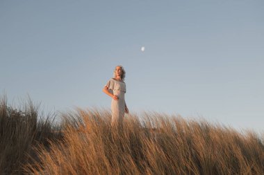 Zarif bir kadın, alacakaranlıkta çimenli bir tarlada durmuş, aya bakıyor. Huzurlu akşam ayarı onun zarafetini ve huzurlu çevresini vurguluyor, doğanın güzelliğini vurguluyor.