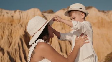 Mutlu bir anne, genç oğluna, güneşli bir çölde şapkasıyla oynarken gülümser. İkisi de beyaz giyinmiş ve birlikte vakit geçiriyorlar..