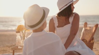 Bir anne ve oğlu, beyazlar içinde, rahatlatıcı bir plaj pikniğinin tadını çıkarırlar. Batan güneş onları mutlu ve kaygısız gösterir..