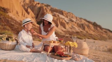 Mutlu bir anne ve oğlu güneşli bir plaj pikniğinin tadını çıkarıyor. İkisi de beyaz giyinmiş meyve ve meyve suyu içiyorlar..