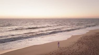 Gün batımında kumsalda ayak izleriyle okyanus dalgalarına doğru yürüyen bir kadının hava aracı görüntüsü..
