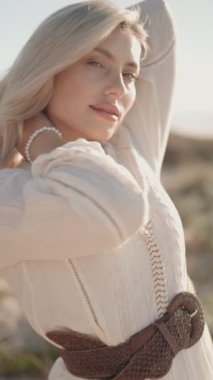 Güzel sarışın kadın dalgalı beyaz bir elbise içinde sahilde poz veriyor. Kameraya gülümsüyor ve güneşli bir günün tadını çıkarıyor..