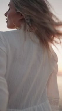 Rüzgâr gibi saçları olan kaygısız bir kadın gün batımında okyanusa doğru yürürken kameraya neşe içinde gülümsüyor..