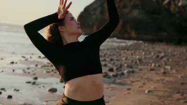 Gün batımında kayalık bir plajda zarif bir şekilde poz veren bir dansçı doğadaki güzelliği ve hareket özgürlüğünü yakalar. Dans, doğa ve sükunet temaları için mükemmel..