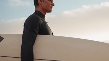 Siyah dalgıç kıyafetli bir sörfçü deniz kıyısına bakarken kolunun altında sörf tahtasıyla yürür..