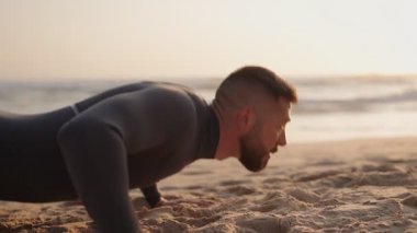 Dalgıç kıyafetli genç bir adam kumlu bir plajda şınav çekiyor. Altın saat boyunca arka planda dalgalar çarpıyor..