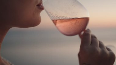 Gün batımında deniz kenarında bir bardak gül şarabı içen bir kadın. Bardak terliyor ve içecekteki kabarcıkları görebilirsiniz. Yavaş çekim, rahatlatıcı, romantik ruh hali..