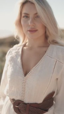 Kahverengi deri kemerli beyaz elbiseli güzel sarışın bir kadın mutlu bir şekilde gülümsüyor ve kameraya bakıyor. Sahilde duruyor, güneş sıcacık parlıyor..