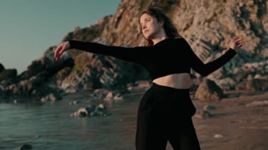 Yetenekli bir dansçı, doğada özgürlük ve zarafeti somutlaştıran, gün batımında kayalık bir plaj arka planına karşı akışkan hareketlerle sanatsal ifadeler sergiler..