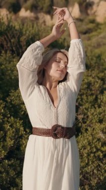 Beyaz elbiseli güzel genç bir kadın yeşil çalıların ve kuru çöl tepelerinin arasında duruyor. Elleri saçlarında, gözleri kapalı ve gülümsüyor..