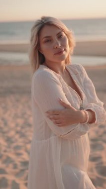 Beyaz elbiseli güzel sarışın hamile kadın, kumsalda altın saat gün batımında gülümseyen, kucaklayan bebek tümseği için poz veriyor. Annelik, hamilelik, beklenti kavramları.