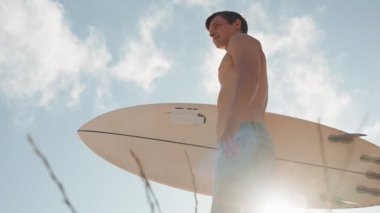 Genç, formda, atletik sörfçü denize bakıyor, sörf tahtasını parlak mavi bir gökyüzünün altında tutuyor. Dalgaları yakalamak için hazır..