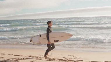 Dalgıç kıyafetli genç bir adam sörf tahtası kolunun altında sahilde koşuyor. Güneşli bir günde sörf yapmaya gittiği için mutlu..