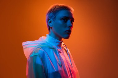 Canlı ışıklandırmayla kapüşonlu bir kıyafet giyen, modern ve sanatsal bir görsel stil sergileyen, koyu renklere sahip ve turuncu gradyan arka plana karşı çarpıcı bir kontrastı olan bir kişinin çağdaş portresi..