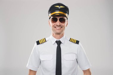 Havacı güneş gözlüğü takan profesyonel bir uçak kaptanı ve standart bir üniforması var. Basit bir gri arka plan önünde kendine güven ve uzmanlık yayıyor. Havacılık alanında profesyonellik ve otorite gösteriyor.