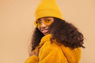 Kendine güvenen, parlak sarı şapkalı, son moda güneş gözlüklü ve tüylü bir ceket giyen genç bir kadın, sıcak tek renkli bej ayarı tamamlıyor..