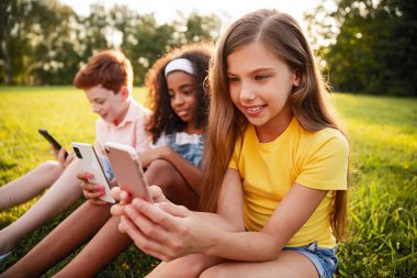 Güneşli bir günde üç çocuk parka yakın otururlar, neşeli bir şekilde akıllı telefonlarıyla meşgul olurlar..