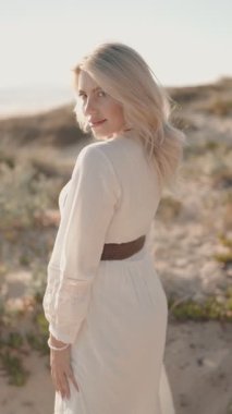 Gün batımında kumsalda yürüyen, beyaz elbiseli ve kameraya gülümseyen, mavi gözlü, çarpıcı sarışın bir kadın..