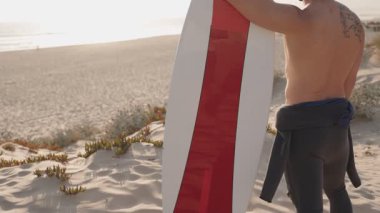 Güneşli bir günde bir sörfçü sörf tahtasıyla sahil boyunca yürür. Dalgıç kıyafeti giyiyor ve dalgaları yakalamaya hazır..