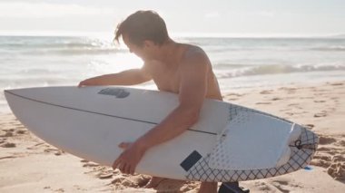 Bir sörfçü güneşli bir sahilde duruyor ve suya girmeden önce sörf tahtasını hazırlıyor. Dalgalarda sörf yapmak için heyecanlı..