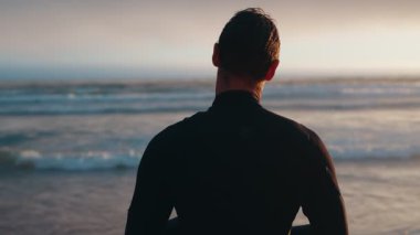 Dalgıç kıyafetli bir sörfçü gün batımında okyanusa bakar ve dalgaları izlerken boynunu ısıtır..
