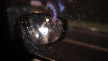 Yağmurlu bir gece görüntüsünü araba dikiz aynasından yakından çek. Güzel trafik ışıkları, ıslak camlardaki yansımalar ve kasvetli şehir atmosferi.. 