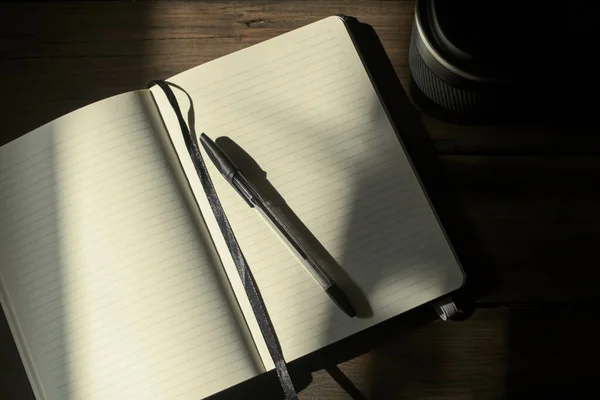 Notebook for notes with a black pen on a wooden table, next to a cup of black color. Photos with sharp contrast and darkening - writing utensils and coffee. Sunlight falls on a notebook with a pen.