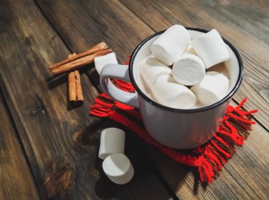 Ahşap masada sıcak çikolata ve marşmelovlu bir kupa var ve yanında da tarçın çubukları var. Noel kompozisyonu - içki, marshmello, tarçın ve örgü bardak standı.