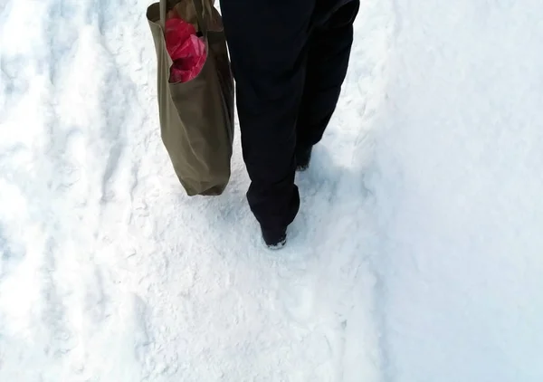 stock image Human legs and shopping bag. a man walks through the snow with a full bag. Bag of groceries - the feet of a man in black pants walk through the snow.