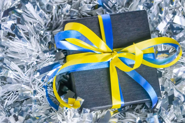 In blue tinsel is a gift with a yellow-blue ribbon. Christmas and New Year - a gift of the symbol of Ukraine. The black gift is tied with a yellow-blue ribbon.