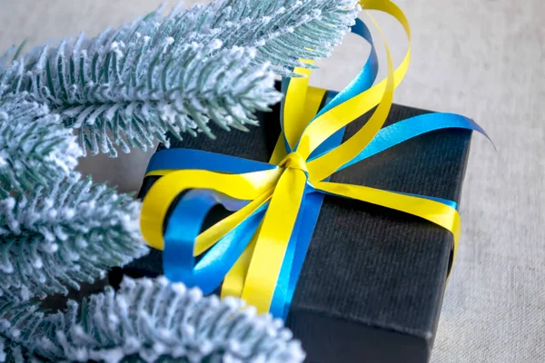 A gift in a black package with a yellow-blue ribbon under the spruce branches. branches of spruce in the snow, a gift with a lazy the color of the flag of Ukraine. Christmas and New Year\' Day.