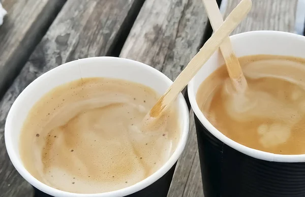 Two cups of coffee on wooden boards. Top view of the coffee cups outside. Paper cups with wooden sticks and frothy coffee.