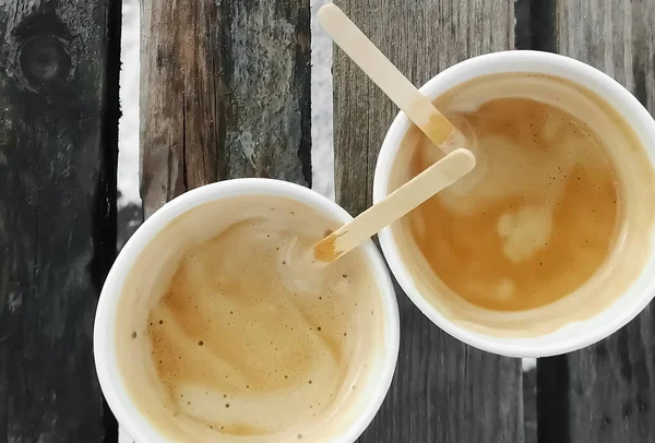 Two cups of coffee on wooden boards. Paper cups with wooden sticks and frothy coffee. Top view of the coffee cups outside.