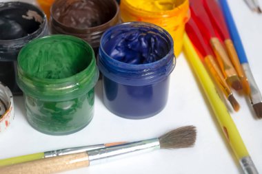 Jars of colored paint stand next to the brushes. For artists, there are a lot of paints and brushes. watercolor and oil paints on a white table.