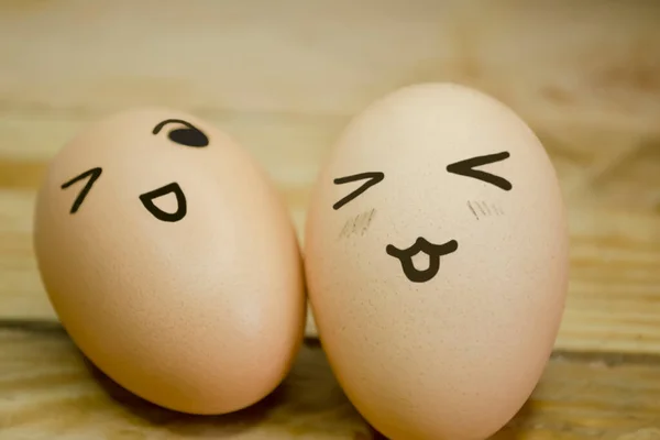 Two eggs with emoticons on the background of a wooden board. On a wooden table are two eggs with emoji. On the eggs, the drawings are emotions. A smile and a wink at the eggs.