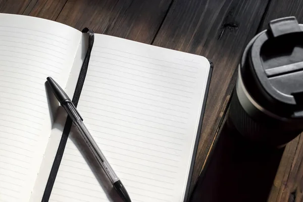 Notebook with a pen on a wooden table next to a cup of coffee. Writing utensils and coffee making facilities. Open notepad with pen.