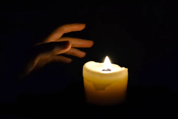 In the dark, a hand reaches for a burning candle. The faint light of the candle illuminates the hand. Hand and candle in a dark room. The warmth of the candle warms the skin.