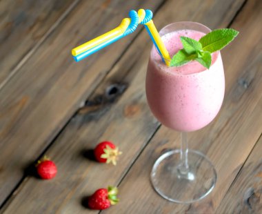 Kamış ve nane yapraklı bir bardakta çilek kokteyli. Ahşap bir masanın üzerinde çilek kokteyli ve nane yapraklı bir bardak var. Bir kokteyl için sarı-mavi pipet.