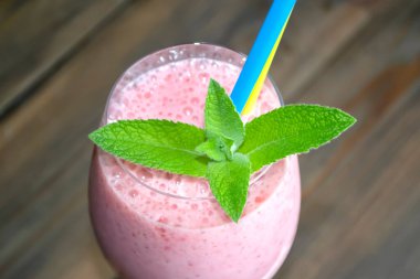 Kamış ve nane yapraklı bir bardakta çilek kokteyli. Kokteyl için sarı-mavi pipet. Ahşap bir masanın üzerinde çilek kokteyli ve nane yapraklı bir bardak var..