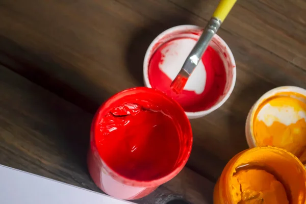 Red paint in a jar on a wooden table. The brush lies on the hook from the jar of paint. Bright colors on the table. Gouache is red.