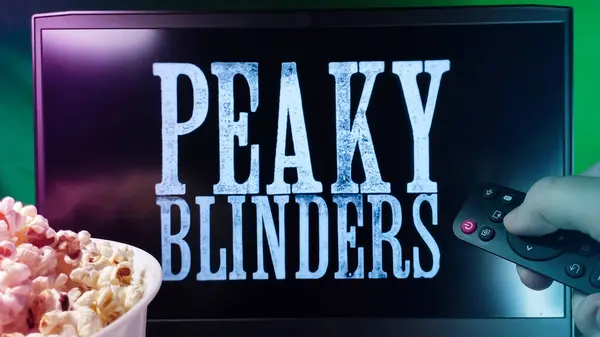 Kyiv. Ukrayna. 10.11.2023: Patlamış mısır ve uzaktan kumandalı Peaky Blinders serisini izleyen kişi. Stok editör fotoğrafı.