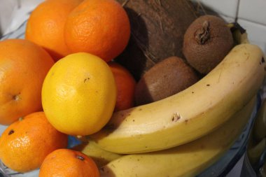 A vibrant assortment of tropical fruits sits in a bowl on a kitchen countertop, featuring yellow bananas, bright oranges, green kiwis, and a large coconut. clipart