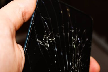 Bir el akıllı telefonu kırık bir ekranla kavrayıp loş bir çevredeki hasarın boyutunu vurguluyor..