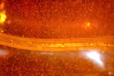 Bir turuncu sıvı şeffaf bir kabı doldurur, kabarcıklar yükselir ve ışık yansımaları canlı renkleri artırırken ince bir tüp kısmen batar.